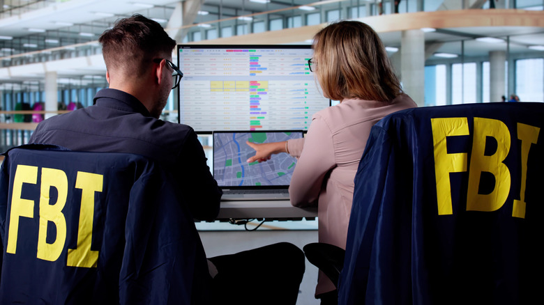 Two people with FBI jackets on the backs of their chairs sit a computer terminal analyzing data