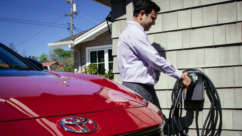 A person standing next to the home wall-mounted charger for their red Toyota electric vehicle.