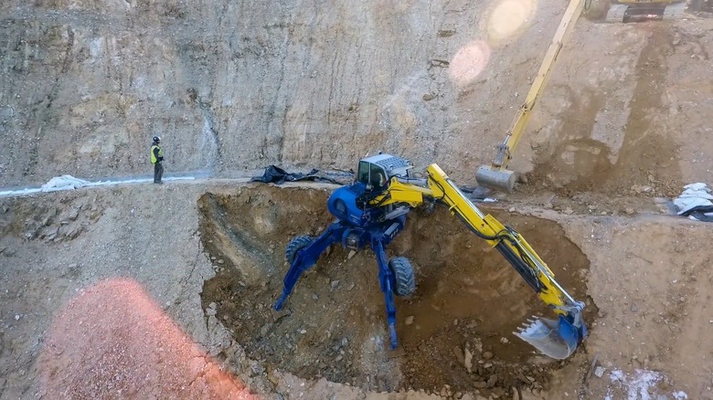 A walking excavator with its stabilizing claws extended, clinging to the side of a rock quarry as it works