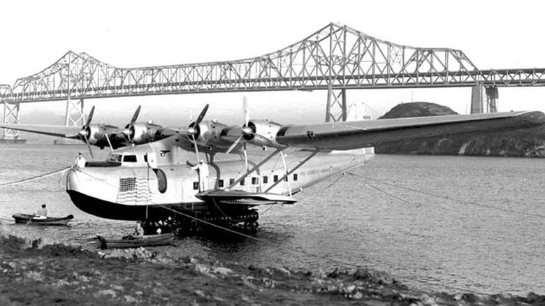 Black and white photo of a large multi-engined seaplane on water near a bridge.