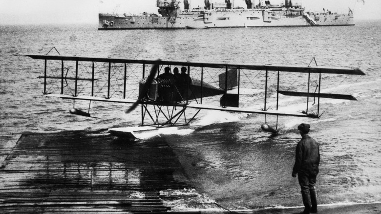 Black and white photo of a small seaplane on water approaching a pier.