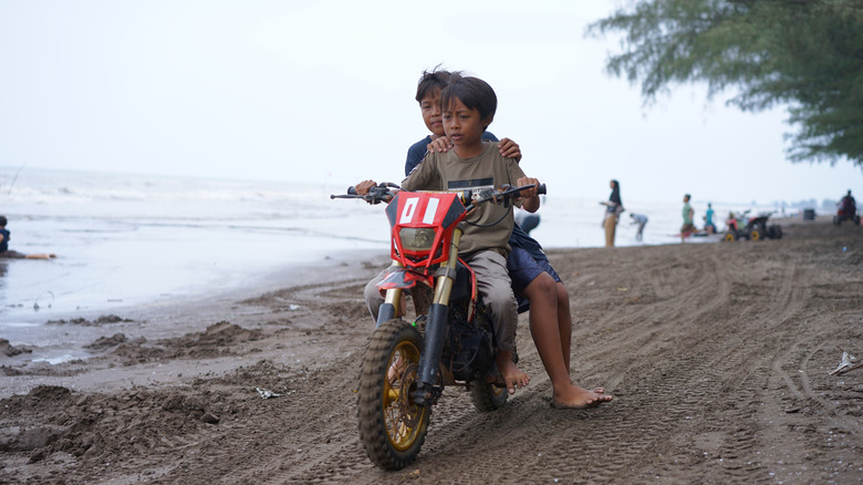 Two kids riding a mini dirt bike on a sandy trail by the shores