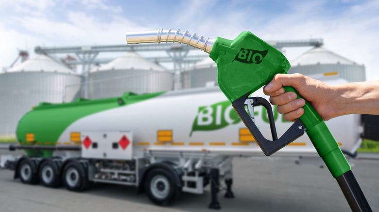 A hand holding a green fuel nozzle with a tanker truck in the back with the word 