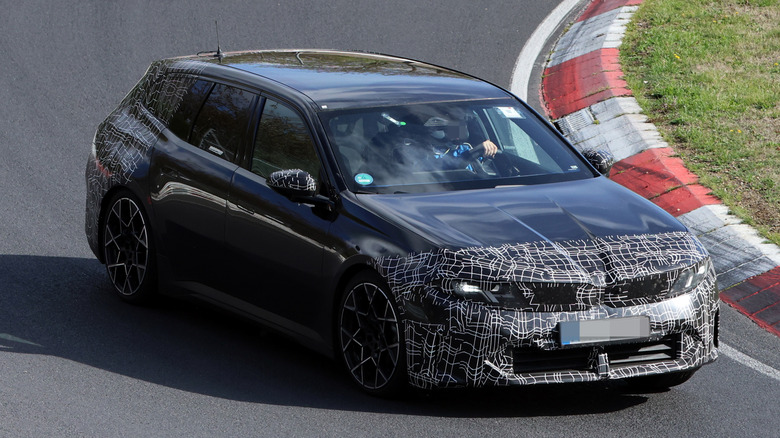 A front three-quarter view of a black camouflaged BMW i3 Touring driving on a track.