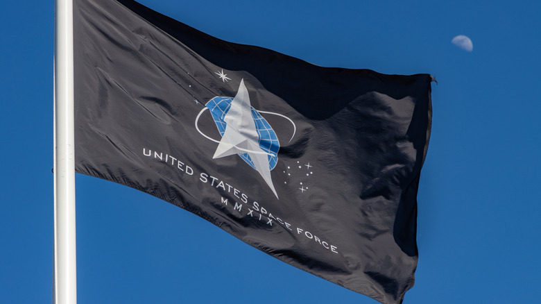 The United States Space Force flag flying against a bright blue sky with the moon visible in the background