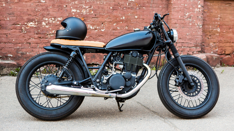 The side view of an unbranded cafe racer motorcycle, black with a tan seat, black helmet on the seat