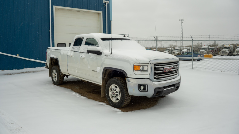 A white GMC Sierra parked outside a warehouse in the snow, front 3/4 view