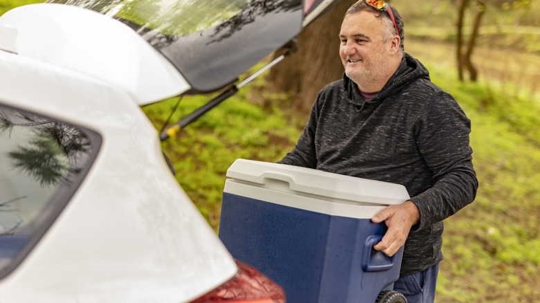 A man in a black hoodie loading a blue cooler to the trunk of a white car