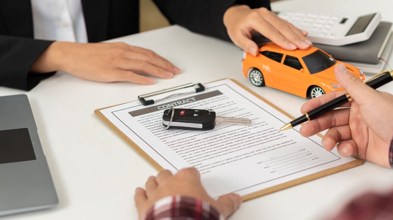 A pair of hands signing a car title document on a desk with a dealer sitting on the other hand