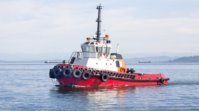 A red tugboat with a white wheelhouse sailing along the hazy hilly coast in the early morning