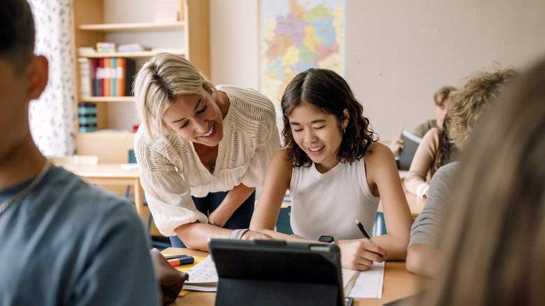 A female teacher leans forward to help student with an activity displayed on an iPad above the student's desk.