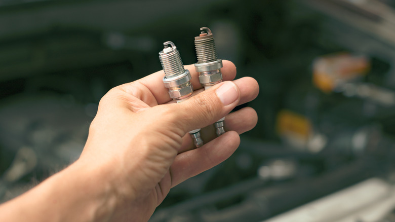 A mechanic holding a new and a used spark plug above the engine bay.