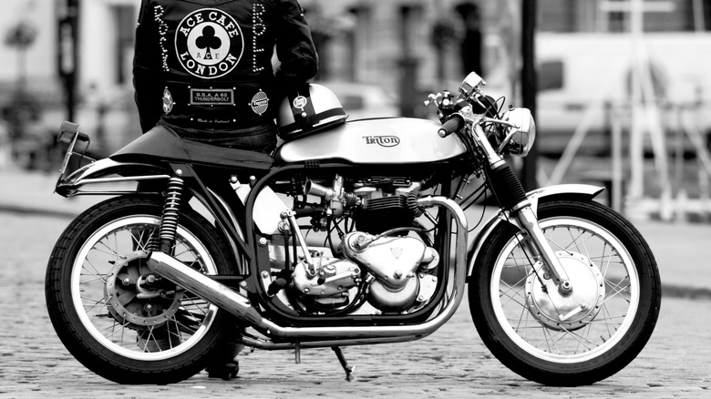 Side view of a motorcycle in a cobbled street on its stand, with a rider leaning against the seat, back to the viewer, in a leather jacked bearing the logo Ace Cafe London.