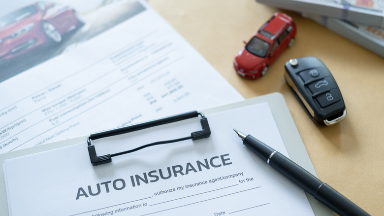 Auto insurance paperwork on a clipboard with a pen, key fob, a red toy car, and a pile of cash sitting nearby