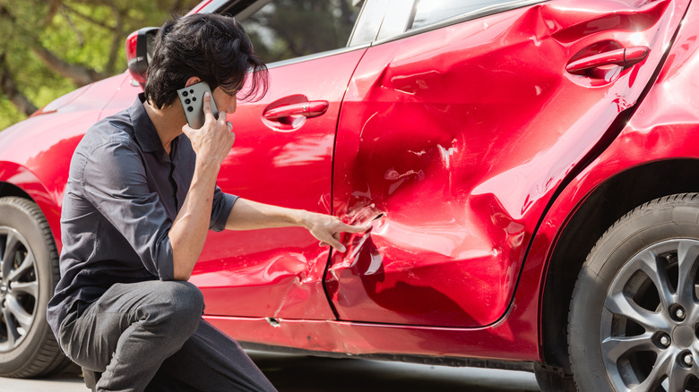 A woman kneeling next to a damaged red car with her cell phone up to her ear