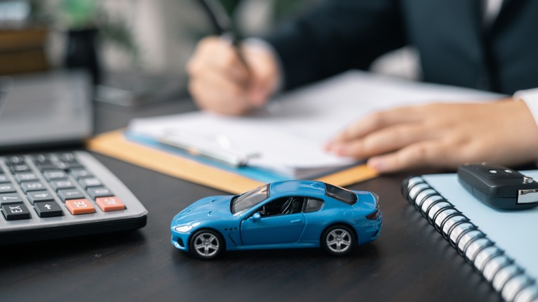 A small blue model car sits on an office table next to a calculator, car keys, and a person filling out a form.