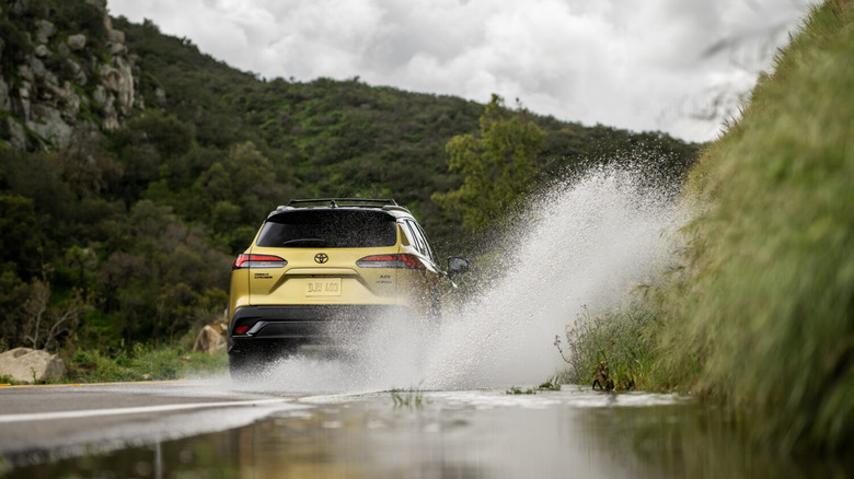 A back end shot of a Toyota Corolla Cross driving though a puddle spraying up water on a mountain road.
