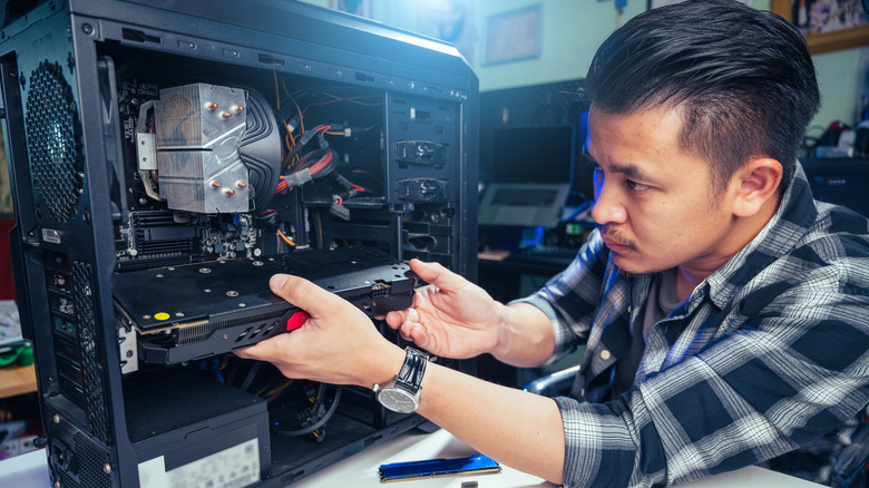 A man with dark hair and a check shirt is removing components from a desktop PC tower that has its side panel removed.