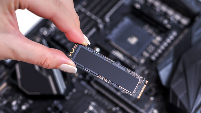 A woman's hand holding an SSD m2 with a computer motherboard in the background.