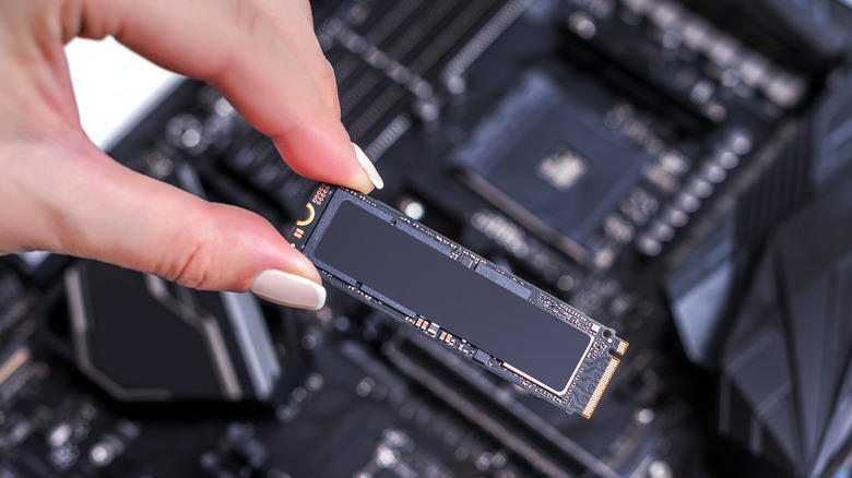 A woman's hand holding an SSD m2 with a computer motherboard in the background.