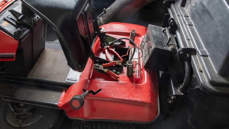A riding lawn mower with its seat lifted revealing a battery and jumper cables