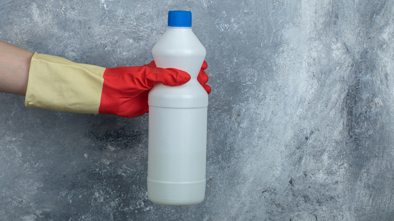 A gloved hand holding an unmarked bleach bottle.