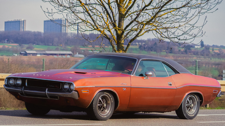 Orange 1970 Dodge Challenger RT parked on road with trees, field, and city in background