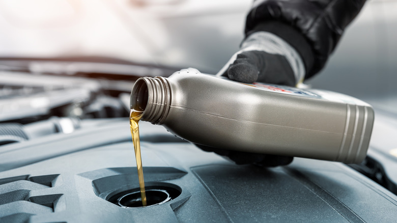 gloved hand holding small bottle can pouring motor oil in car engine