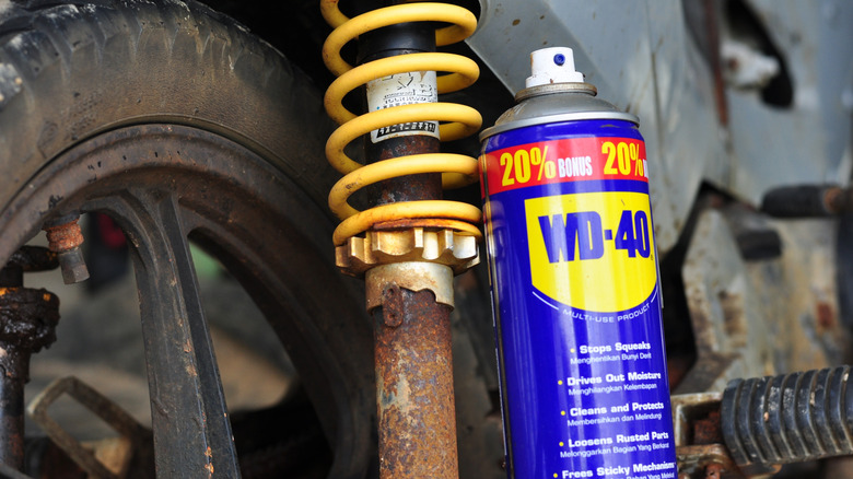 A bottle of WD-40 next to a rusty shock absorber with a yellow coil spring, dirty wheel and tire next to it