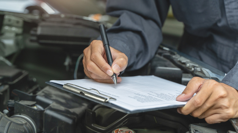 A mechanic fills out form on clipboard above car engine