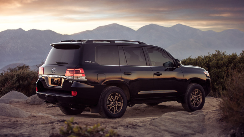 A black Toyota Land Cruiser heritage edition parked at the edge of a dirt area with foliage in the back.