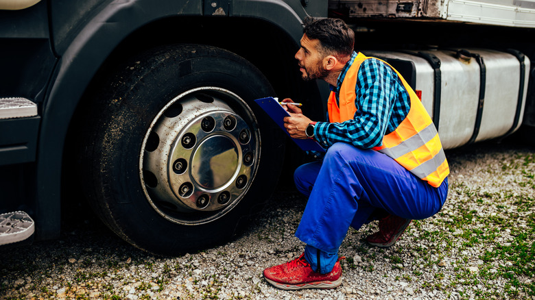 Truck driver crouching next to the front wheel of his semi-truck, performing an inspection and taking notes on a clipboard