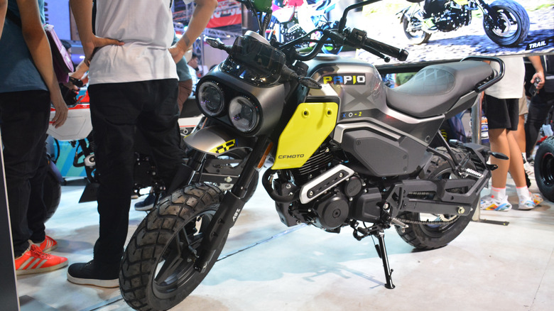 A black CFMoto Papio motorcycle on display at an indoor venue trade show, standing on its kickstand.