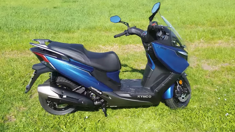 A dark blue Kymco X-Town 300 scooter motorbike parked on grass, facing towards the right.