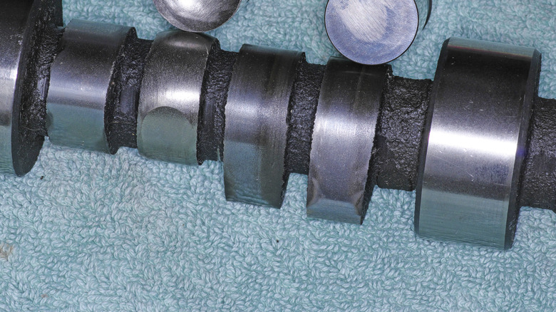 A camshaft sitting on a table with two lifters, with the lifter on the left cupped and the matching camshaft lobe eaten away