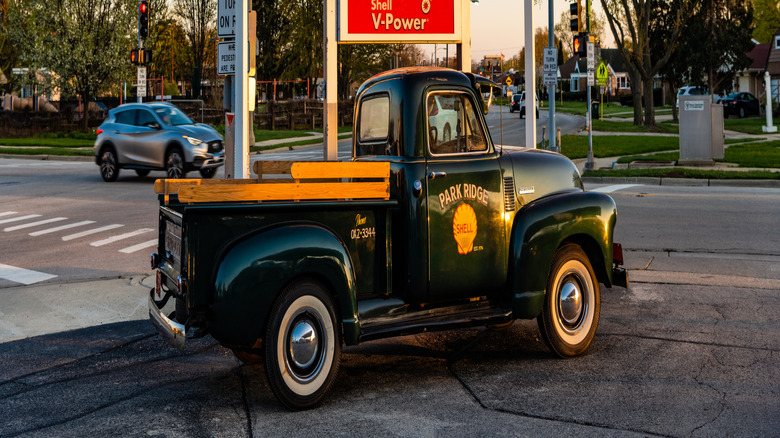 Antique black Chevrolet truck with whitewall wheels sitting in front of a Shell gas station sign with a price of 4.14 per gallon for regular