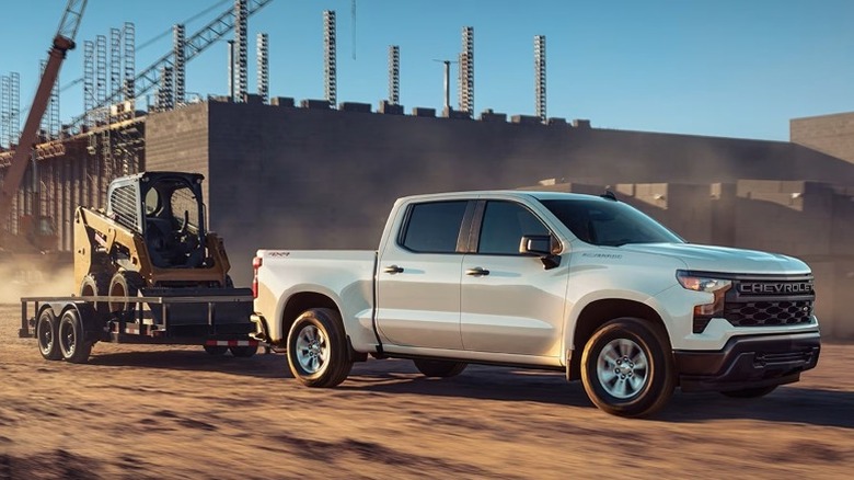 Chevy Silverado 1500 WT towing a flatbed trailer carrying a skid-steer loader at a construction site