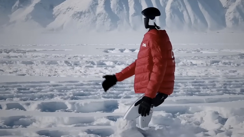 A Unitree G1 robot in an orange puffer jacket walking in knee-deep snow
