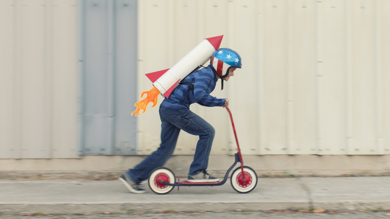 Boy on scooter wearing cartoonish helmet with a rocket strapped to his back
