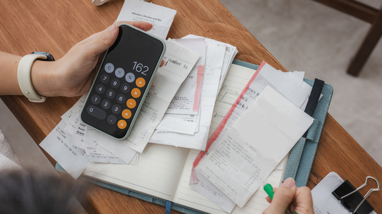 A person using a calculator app on their phone and several paper receipts to balance their budget
