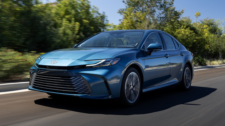 A blue 2025 Toyota Camry driven down the road with green trees in the background.
