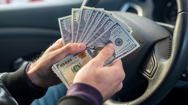 Person fanning out multiple $100 bills while seated at the wheel of a car.