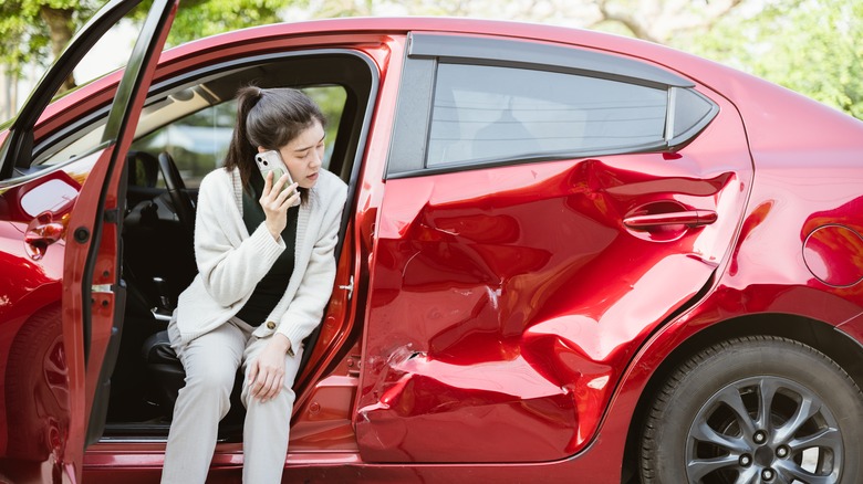 woman on cell phone looking at dented car after crash