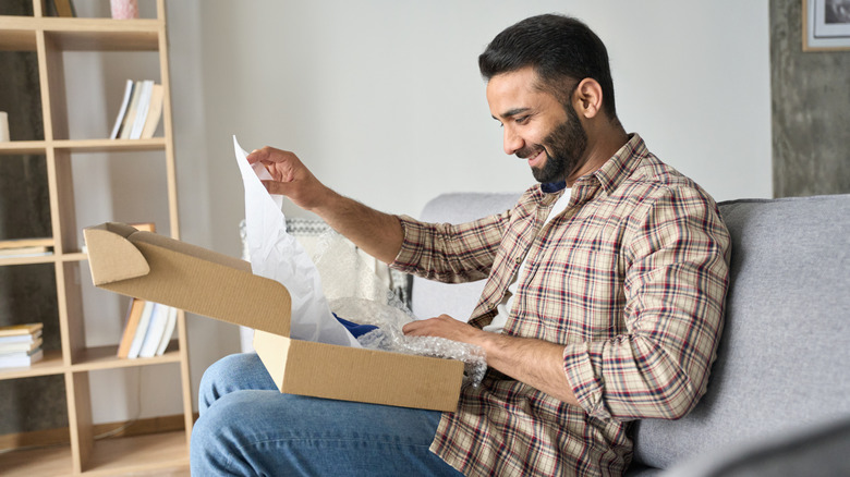 A bearded man wearing plaid shirt and denim jeans sits on the couch to unwrap a present