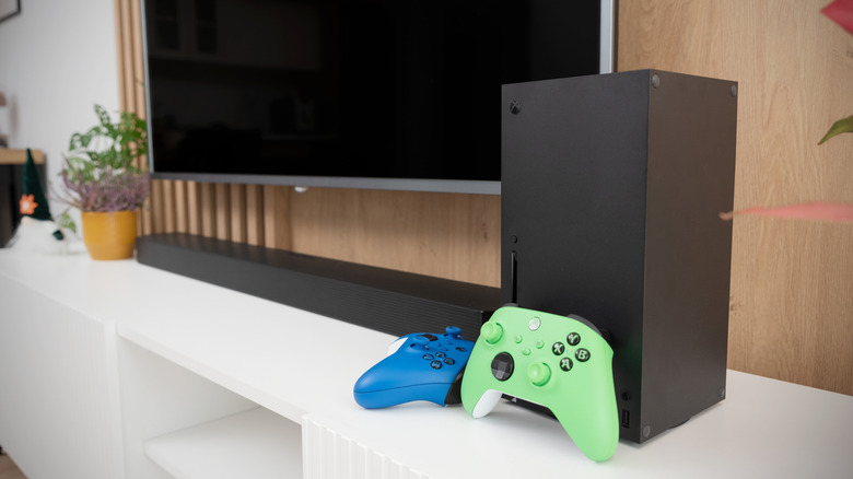 An Xbox Series X in black, accompanied by two controllers, sits on a TV stand with a television in the background.