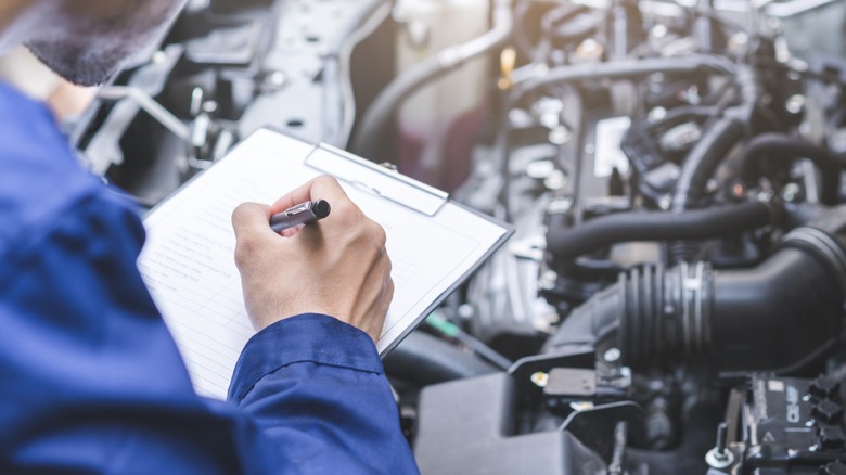An auto mechanic writing on a clipboard in front of an engine