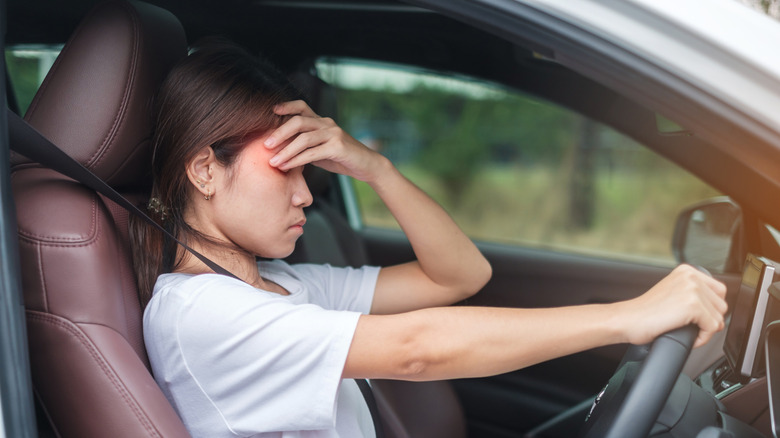 A woman seated in a car with her eyes closed and her hand to her forehead, appearing stressed