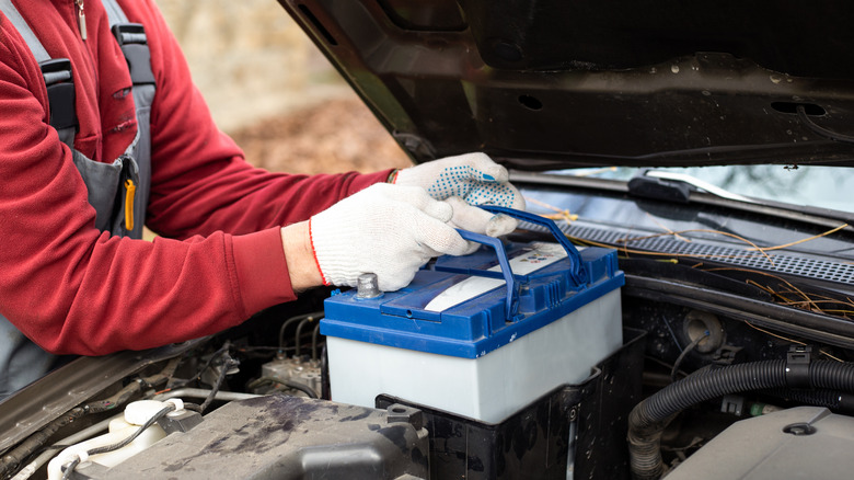 A close up of a mechanic in gray overalls and a red shirt lifting out a battery from a car