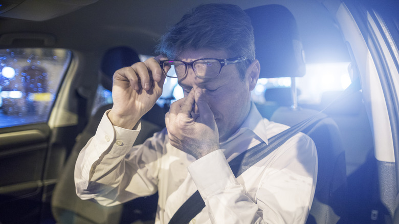 A driver lifting his glasses and pinching the bridge of his nose due to bright lights behind him