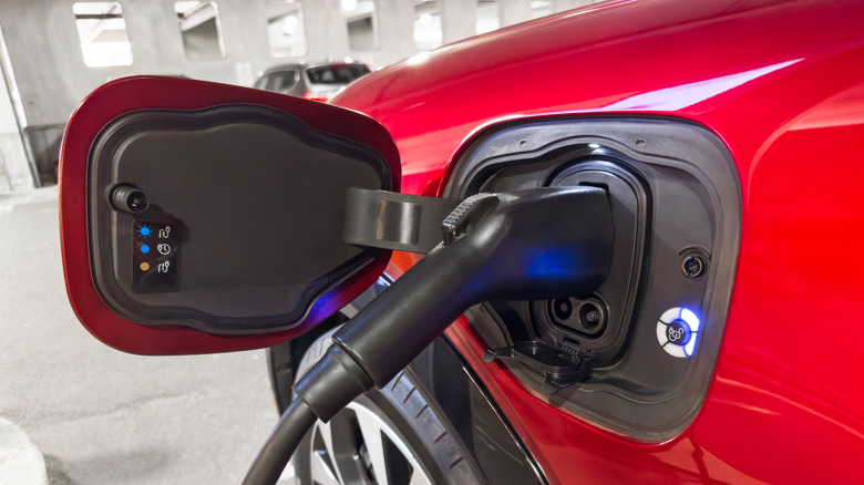 A close-up view of an electric vehicle charging plug connected to the charging port of a red car in a parking garage, with a glowing blue indicator light visible around the port signaling an active charge